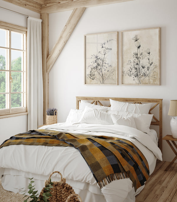 A pair of framed botanical prints featuring delicate black roses and other flowers in shades of grey and white hang above a cozy bed in a rustic bedroom with exposed wooden beams.