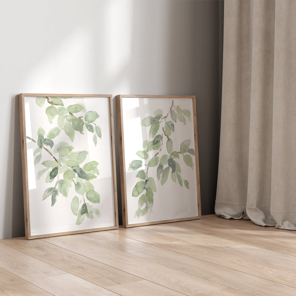 Two watercolor leaf prints in oak frames leaning on a light wood floor