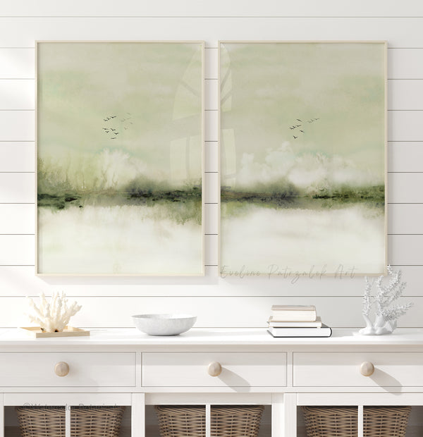 A pair of vertical framed abstract wall art prints with soft sage green, cream, and gray tones and flying black birds, displayed side-by-side on a white shiplap wall above a white console table with decorative coral and a bowl.