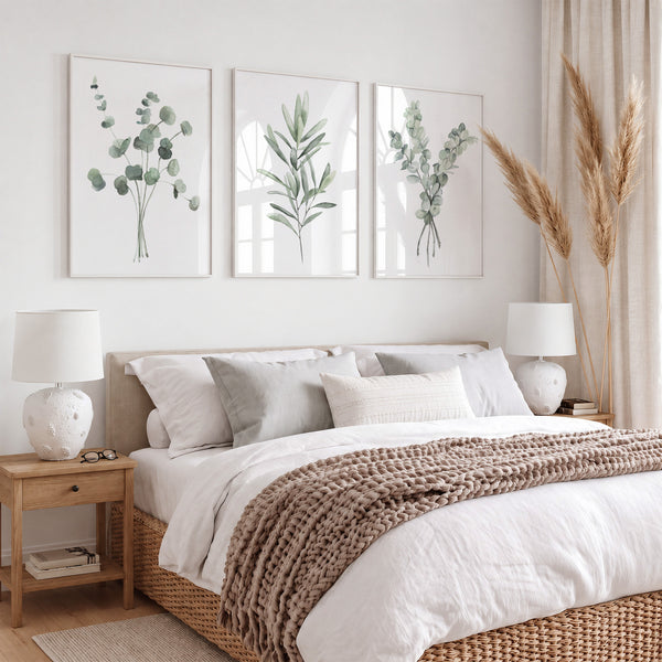 A styled bedroom scene with a bed dressed in white bedding and a chunky brown knit throw, beneath three framed botanical prints in sage green on a light wall. The room includes wooden bedside tables with white lamps, beige curtains, and dried pampas grass.
