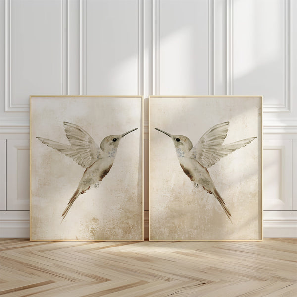 set of 2 prints standing on the floora nd leaning against white panelled wall in wooden frames showing 2 hummingbirds facing each other