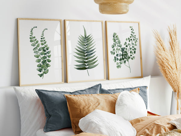 Three rectangular, light wood-framed prints featuring detailed watercolor illustrations of different fern varieties are displayed above a white upholstered headboard in a bedroom. The bed is made with a beige duvet and has several pillows in shades of blue, brown, and white. A large, tan woven pendant light hangs above the bed. On the right, a tall vase holds feathery pampas grass.