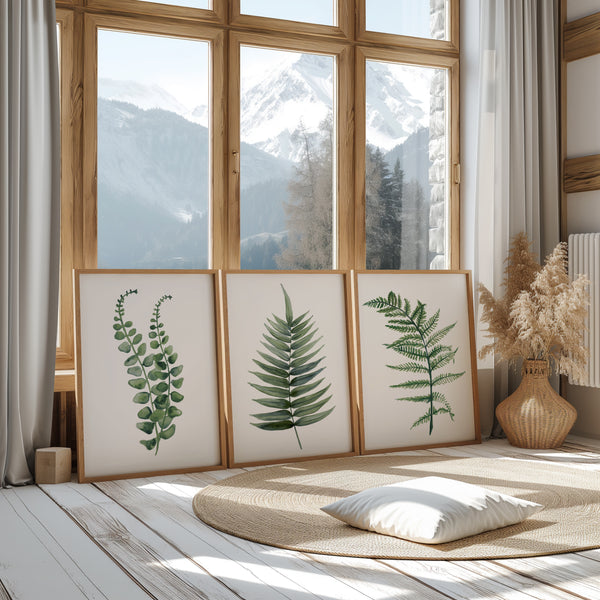 3 Holzrahmen mit minimalistischen grünen Farnblättern stehen auf dem Boden und lehnen sich an ein großes Fenster mit Blick auf die Berge.