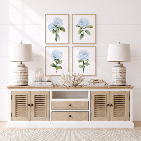 Light wood console table with lamps, candles, and decorative items against a white wall with framed floral art showing minimalist light blue hydrangea prints.