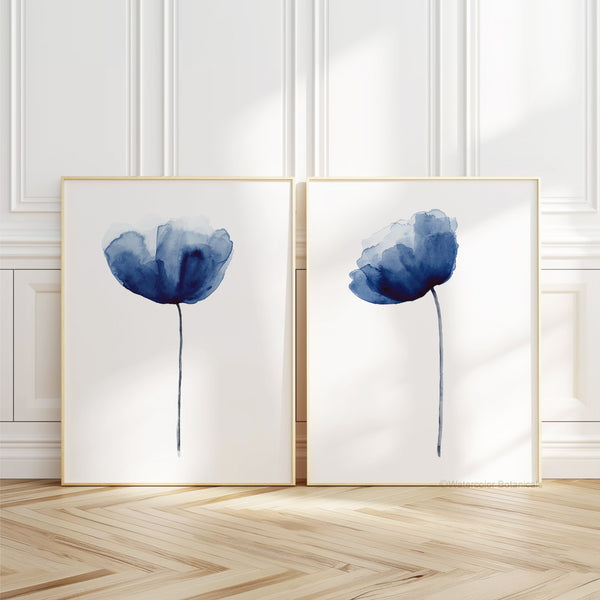 A pair of framed watercolor paintings depicting elegant navy blue poppies, displayed on a white wall with ornate molding. The paintings are framed in a simple, natural wood frame, complementing the clean lines of the wall decor. The overall aesthetic is minimalist and modern, with the poppy prints adding a touch of color and visual interest.