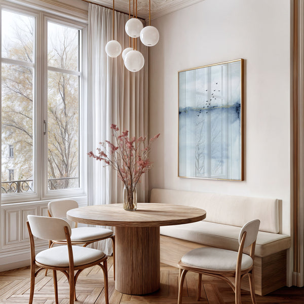 A framed Blue Watercolor Abstract painting of a blue and white landscape with a flock of birds flying across the sky hangs above a round dining table in a bright and airy dining room.