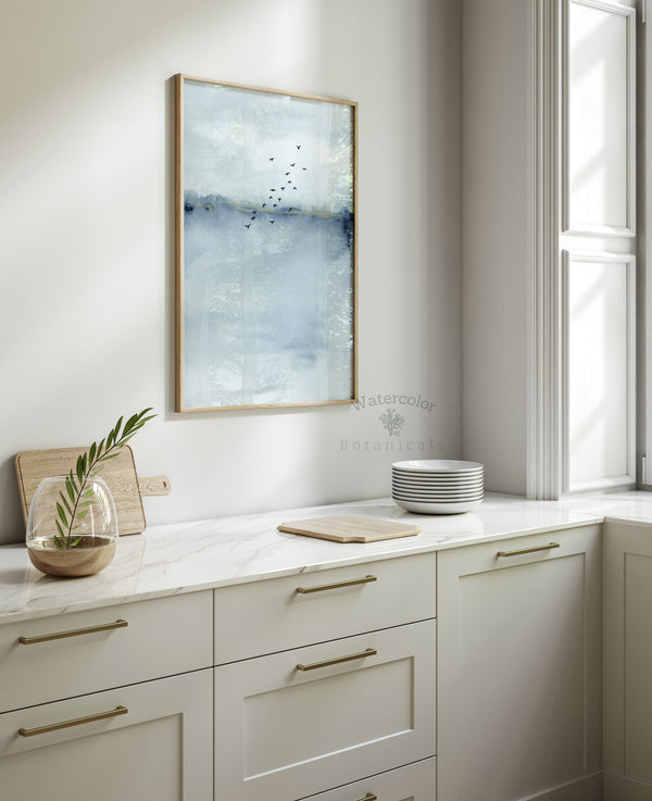 A framed Blue Watercolor Abstract painting of a blue and white landscape with a flock of birds flying across the sky hangs in a kitchen above a white countertop.