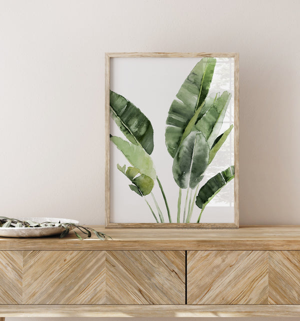 A watercolor painting of large green tropical banana leaves adds a touch of nature to a living room with a wooden dresser and a white wall.