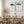 Mediterranean art is showcased with two framed watercolor illustrations of Italian Stone Pines hanging on a textured white wall in a dining room. The trees are depicted with green foliage and brown trunks, each in a thin black frame. Below the artwork, a long, oval wooden dining table is set with several matching wooden chairs. Two ceramic vases, one holding dried grasses, sit on the table. The room is bright, with a sheer white curtain partially visible on the left.