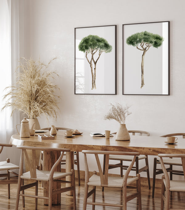 Mediterranean art is showcased with two framed watercolor illustrations of Italian Stone Pines hanging on a textured white wall in a dining room. The trees are depicted with green foliage and brown trunks, each in a thin black frame. Below the artwork, a long, oval wooden dining table is set with several matching wooden chairs. Two ceramic vases, one holding dried grasses, sit on the table. The room is bright, with a sheer white curtain partially visible on the left.