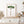 Mediterranean art is showcased with a framed watercolor illustration of an Italian Pine Tree hanging on a white wall above a light-colored wooden cabinet. The tree is depicted with green foliage and a brown trunk, framed in a thin light wood frame. On the cabinet, there is a white vase holding dried flowers, a set of salt and pepper shakers, and a small decorative object. A portion of a wooden table and a glass vase with branches are visible in the foreground. The scene is bright and natural.