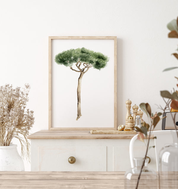 Mediterranean art is showcased with a framed watercolor illustration of an Italian Pine Tree hanging on a white wall above a light-colored wooden cabinet. The tree is depicted with green foliage and a brown trunk, framed in a thin light wood frame. On the cabinet, there is a white vase holding dried flowers, a set of salt and pepper shakers, and a small decorative object. A portion of a wooden table and a glass vase with branches are visible in the foreground. The scene is bright and natural.