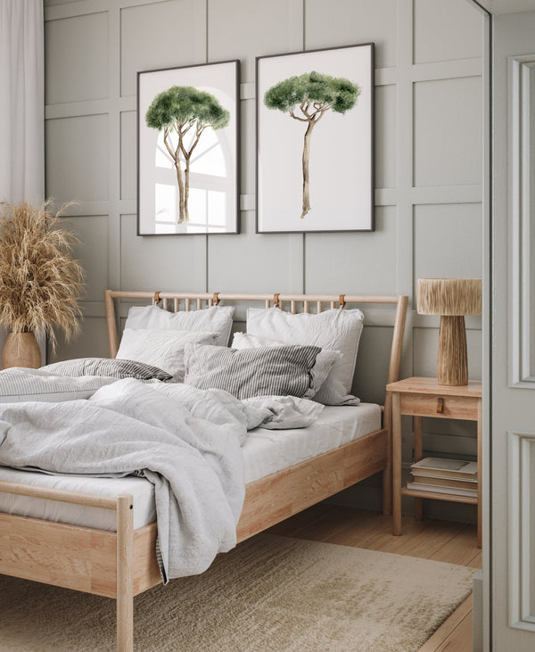 Mediterranean art is featured in a bedroom with two framed watercolor illustrations of Italian Stone Pines hanging on a gray paneled wall above a wooden bed frame with light-colored bedding. The trees are depicted with green foliage and brown trunks, each in a thin black frame. To the left, a woven vase holding dried grasses sits on the floor. To the right, a wooden nightstand with a woven lamp is visible. The bedroom has a light-colored rug and a partially visible white door with paneling.