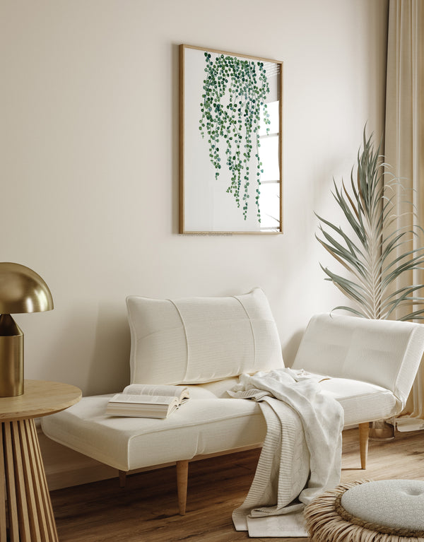 A watercolor painting of a hanging plant with green tropical leaves adds a touch of nature to a living room with a white daybed, a round side table, and a gold lamp.