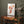 A framed watercolor painting of a ginkgo leaf branch. The delicate, fan-shaped leaves are depicted in warm autumn hues against a white background. The painting hangs on a textured, gray wall above a wooden console table.