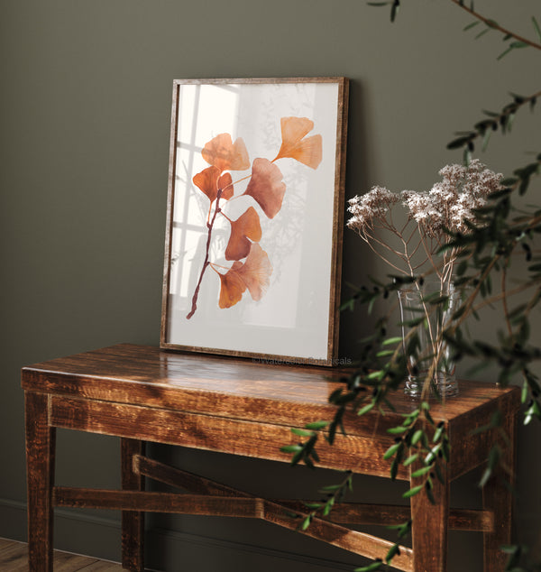 A framed watercolor painting of a ginkgo leaf branch. The delicate, fan-shaped leaves are depicted in warm autumn hues against a white background. The painting hangs on a textured, gray wall above a wooden console table.