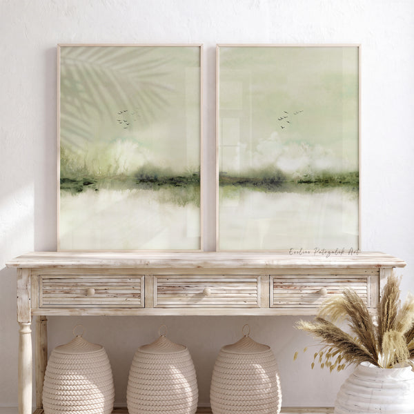 A pair of framed abstract landscape art prints in sage green and cream tones, displayed side-by-side on a distressed white console table in a bright, modern room.