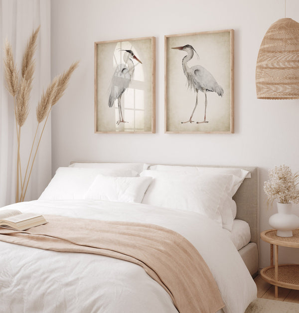 A pair of framed watercolor paintings depicting elegant great blue herons in soft neutral tones against a vintage-inspired background. The paintings are displayed on a white wall above a bed in a bedroom with a serene and calming atmosphere.