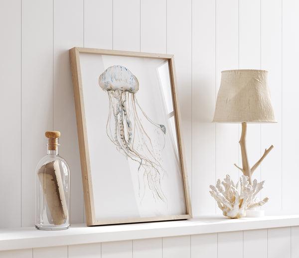 Neutral watercolor jellyfish print in a light wood frame on a white shelf with a driftwood lamp