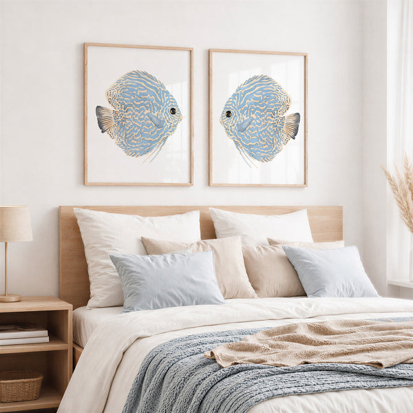 A styled bedroom scene with two framed fish wall art prints above a bed. The prints show blue and gold striped fish on white backgrounds, hung on a light wall above a wooden headboard with white and beige bedding and a blue throw blanket.