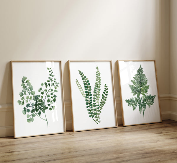 Three green watercolor fern prints in light oak frames leaning against a cream wall on a wooden floor