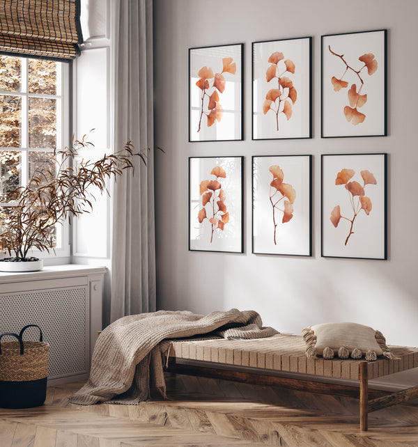 Six framed botanical prints featuring orange-gold ginkgo leaves arranged in two columns on a light gray wall above a wooden bench in a styled room with a window, curtains, and a throw blanket.