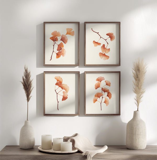 Four square framed pictures on a light wall, each filled with a watercolor painting of ginkgo leaves in warm autumn hues. The delicate leaves are depicted in detail against a white background. In the foreground, two vases with dried pampas grass and white candles on a table create a cozy atmosphere.