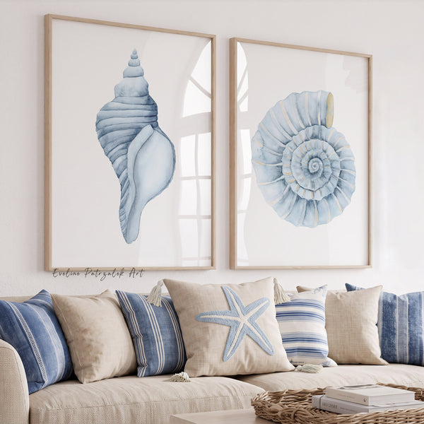 Two framed vertical art prints featuring light blue seashell and nautilus shell illustrations, displayed on a white wall above a beige sofa with blue and beige pillows.