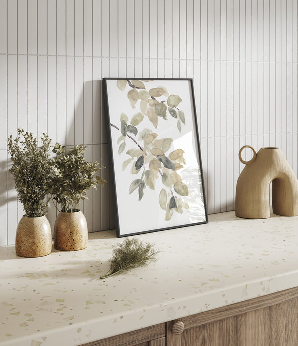 A framed watercolor painting of leaves hangs on a wall above a counter. The counter features a speckled terrazzo surface, and there are small vases with dried flowers and a decorative ceramic vase on the counter.