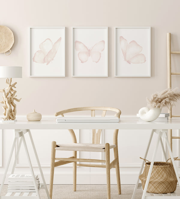 A set of three framed watercolor paintings of delicate pink butterflies, arranged in a row on a light-colored wall above a wooden desk. The desk is adorned with a laptop, a lamp, and various decorative objects, creating a cozy and inviting workspace. The soft pink hues of the butterflies and the overall light and airy aesthetic of the living room create a serene and inspiring atmosphere.