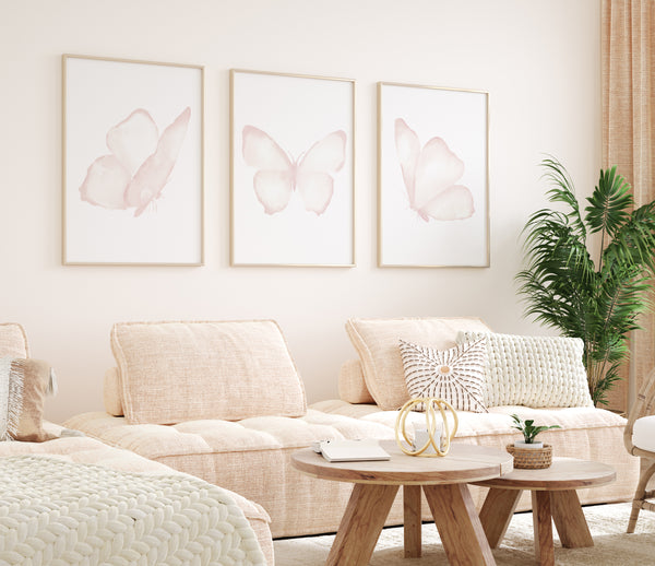 A set of three framed watercolor paintings of delicate pink butterflies, arranged in a row on a light-colored wall above a cozy seating area. The butterflies are depicted with soft, ethereal details, creating a sense of lightness and airiness. The living room is filled with soft textures and natural light, creating a serene and inviting atmosphere. The overall aesthetic is feminine and dreamy, with the butterfly art adding a touch of whimsy and elegance.