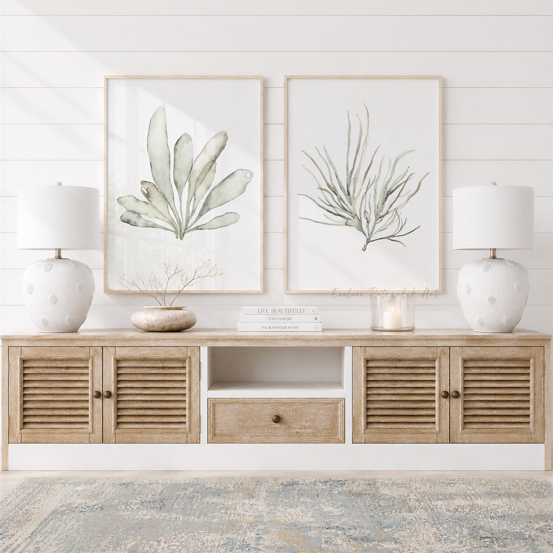 A styled interior scene showing a light wood media console with louvered doors, two white table lamps, and two framed botanical prints on a white shiplap wall.