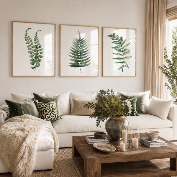 Three framed botanical prints of green ferns hang above a white sectional sofa in a styled living room with green and beige pillows, a wooden coffee table, and a vase of greenery.