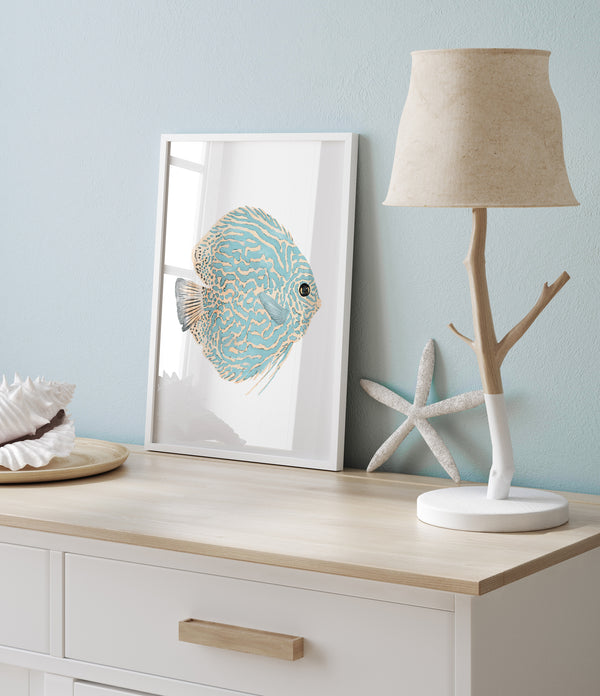 A framed watercolor painting of a discus fish with vibrant blue and yellow patterns. The artwork is displayed on a dresser with a coastal-themed decor, including a lamp and seashells.