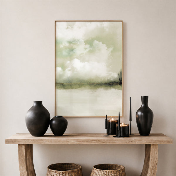 A framed landscape art print featuring soft green and white clouds over a calm body of water, displayed on a light beige wall above a rustic wooden console table styled with matte black vases, candles, and woven baskets.