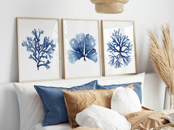 Watercolor paintings of minimalist seaweed and coral designs in deep navy blue add a touch of nature to a bedroom with blue and brown pillows and a white bed.